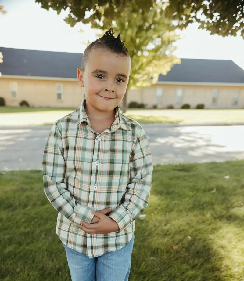 A young Cornerstone Community Schoolhouse student in a plaid shirt standing under a tree outside the school